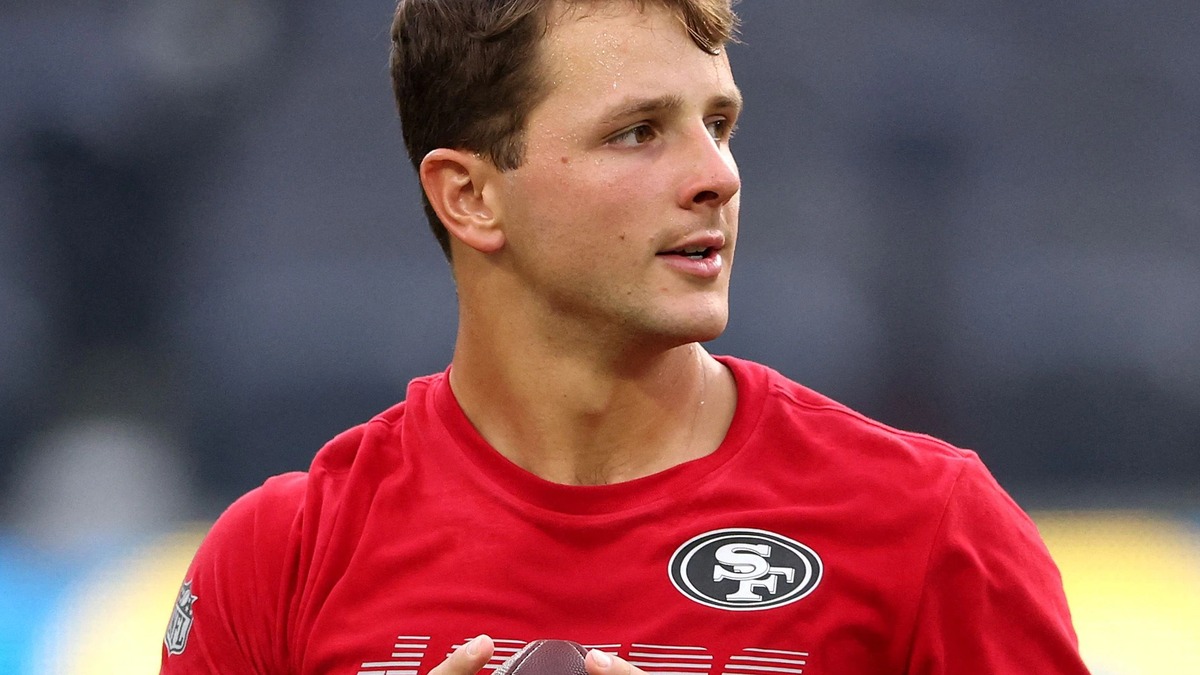 Brock Purdy in a red 49ers warmup shirt holding a football before a preseason game