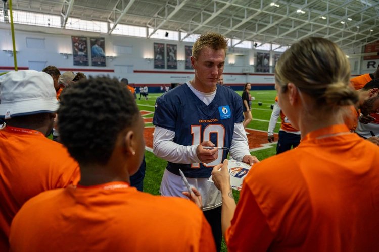 Broncos quarterback Bo Nix in a navy practice jersey signing autographs for young fans at an indoor facility