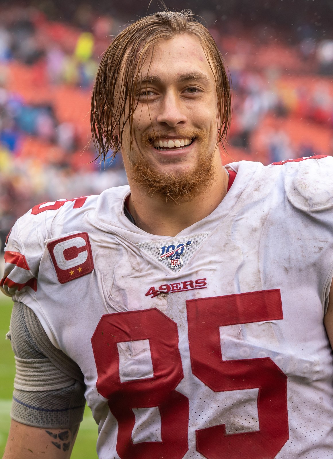 George Kittle smiling in a muddy white 49ers jersey with a captain's patch after a game